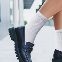 White crew socks with ribbed texture, worn with black loafers, featuring a logo. Suitable for casual or stylish outfits.