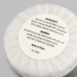 A white circular soap wrapped in paper, labelled with ingredients and warnings, features a logo.