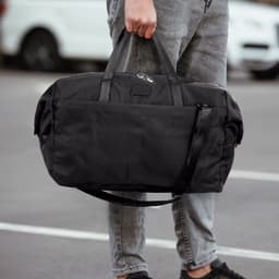 A black duffle bag is being held by a person, featuring dual handles and a shoulder strap.