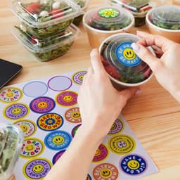 A hand holding a salad container with a glossy sticker. A sticker sheet featuring colourful circular designs is beside it.