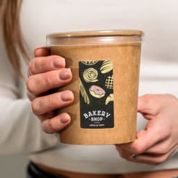 A hand holding a matte rectangular sticker on a brown takeout container, featuring bakery-themed illustrations.