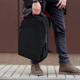 A black daily backpack is being held by a person wearing dark jeans and brown boots, standing on a paved surface.