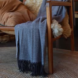 A textured grey blanket drapes over a wooden chair, with fringes at the edge and a soft background.