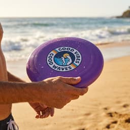A purple frisbee with a logo, held by a person at the beach, with waves and sand in the background.