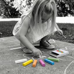 A child uses colourful chalk sticks, including white, orange, pink, green, and purple, from an open box on the ground.