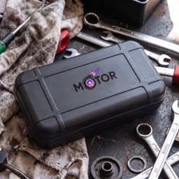 A black tool case sits on a workbench surrounded by various tools and a dirty cloth. It features a logo on top.