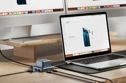 A grey recycled aluminium USB hub with multiple ports, placed on a desk beside a laptop and computer monitors.