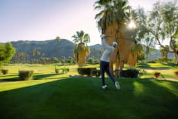 A golfer swings a club on a lush green golf course, surrounded by palm trees and mountains in the background.
