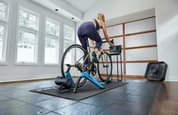 A cyclist using a stationary bike trainer on a mat indoors, with a fan and a display monitor nearby.