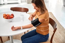 A woman in a brown knitted top uses a black blood pressure monitor on her arm at a table with apples.