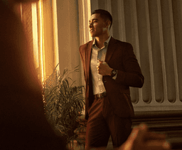 A man in a brown suit stands beside a palm plant, wearing a watch with a soft gold finish and a cream band.