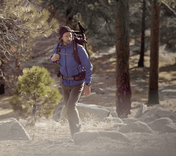 A man wearing a blue jacket and backpack walks through a forested area with rocky terrain.