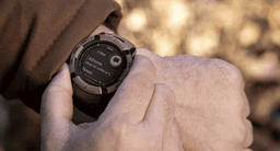 A tactical smartwatch with a rugged design and dark colour, being adjusted on a person's wrist outdoors.