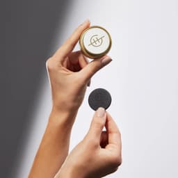 A gold circular container with a logo held in one hand, and a black disc in the other hand.