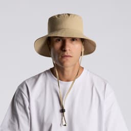 A beige nylon wide brim bucket hat with a drawstring, featuring a logo, worn by a person in a white shirt.