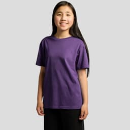 A young girl in a purple t-shirt stands smiling, with long hair and wearing black trousers. The shirt features a logo.