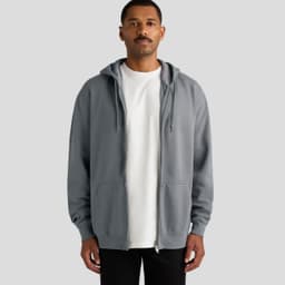 A man stands wearing a grey zip-up hoodie with a hood and kangaroo pockets. The hoodie has a logo on it.