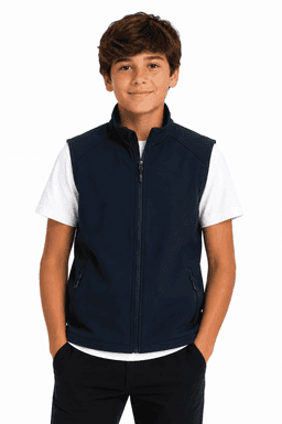 A boy stands wearing a navy blue vest over a white T-shirt, with his hands in his pockets. The vest has branding.