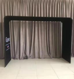 A black fabric arch frame with a tension design, set against a neutral curtain backdrop. Features a logo on one side.