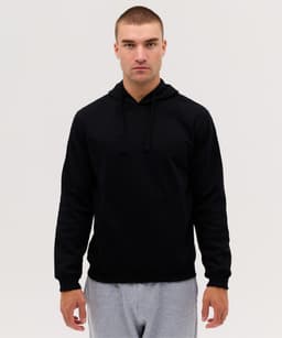 A black hooded jumper with a front pocket and drawstrings, worn by a model. The model is also wearing grey pants.
