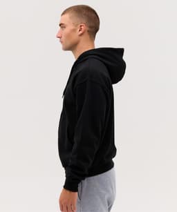 A side view of a man wearing a black zip hoodie with a hood, paired with grey pants. The hoodie has a logo.