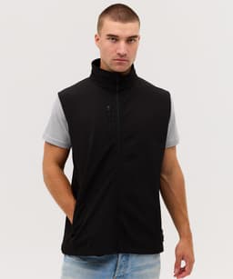 A man in a black softshell vest stands against a plain background, wearing a grey T-shirt and jeans.