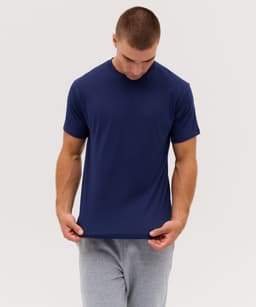 A navy blue t-shirt is worn by a male model, who is examining the hem. The model is also wearing grey trousers.