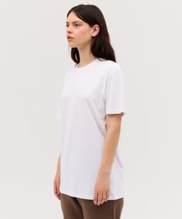 A woman wearing a white T-shirt, standing sideways against a neutral background. The shirt has a simple design and no visible branding.