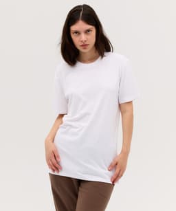A model wears a plain white T-shirt paired with brown trousers, featuring a simple design and a logo.