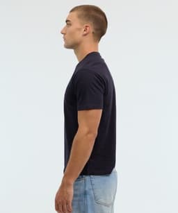 A side view of a man wearing a navy blue essential t-shirt, paired with light blue jeans. The shirt features a logo.