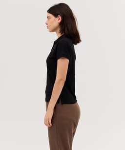 A black women's polo shirt viewed from the side, featuring a collar and a fitted design. It has a logo on it.