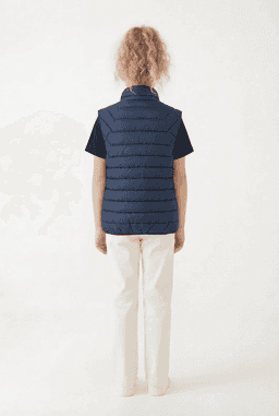 A navy puffer vest viewed from the back, worn over a dark shirt and white trousers. It has a logo on the chest.