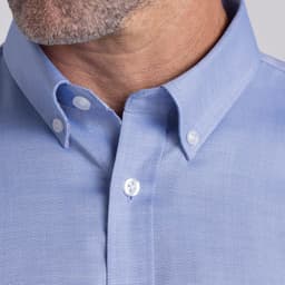 A close-up of a men's button-up shirt in light blue, featuring a pointed collar and buttons. It has a branding logo.