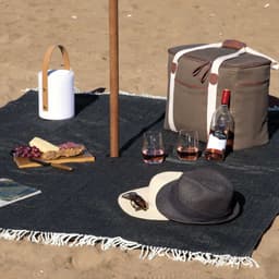 A dark blue picnic blanket on sand with a cooler bag, drink bottle, glasses, snacks, hat, and magazine.