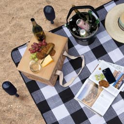 A picnic setup on a checkered rug featuring a timber picnic basket, wine, snacks, and a magazine.