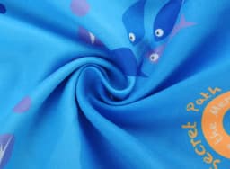 A blue beach towel with a double-faced pile featuring playful fish designs and circular motifs. It has a branding logo.