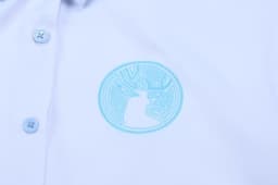 A light blue shirt featuring a circular logo in a similar tone. The shirt has a polo neck and button closure.