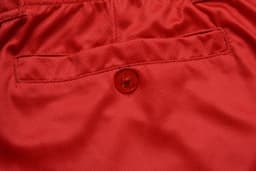 A close-up of red fabric with a button and a pocket detail. The material has a smooth texture.
