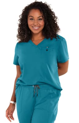 A woman in a teal scrub top with a v-neck and a chest pocket, smiling and standing with hands behind her back.