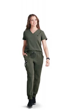 A woman in an olive green scrub top and matching pants, walking confidently and smiling. The outfit has a logo.