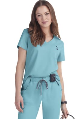 A woman models a teal scrub top with a v-neck and short sleeves. The outfit includes pockets and has a logo.