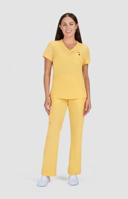 A woman stands in a yellow scrub set, featuring a short-sleeve top and matching trousers. The outfit has a logo.