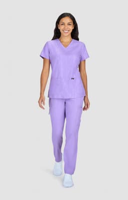 A woman wearing a lavender scrubs set, featuring a V-neck top and matching pants, with a logo visible.