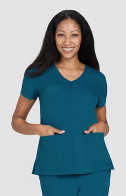 A teal scrub top featuring short sleeves and two front pockets, modelled by a smiling woman.