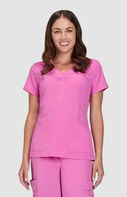 A pink scrubs top featuring a V-neck design and two front pockets, modelled by a smiling woman.