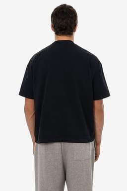 A short sleeve black garment dye t-shirt viewed from the back, worn with grey trousers. The shirt features a logo.