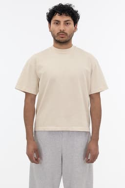 A short sleeve garment dye t-shirt in a beige colour, worn by a model with curly hair and grey pants.