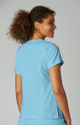 A woman with curly hair wearing a light blue top, featuring a logo on the sleeve, facing away from the camera.