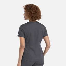 A woman with curly hair wearing a grey tuckable V-neck top, viewed from the back. The top has a logo on the sleeve.
