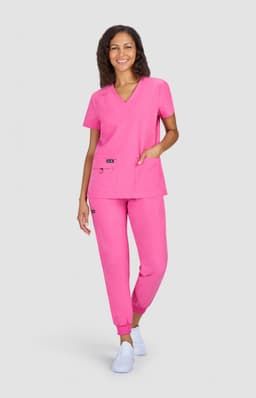 A woman stands in a pink scrub top and matching pants, featuring pockets and a logo on the top.